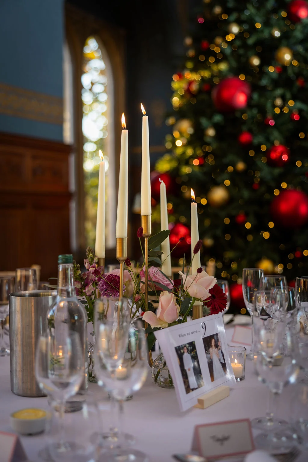 An elegantly styled wedding reception table featuring tall taper candles, soft pink floral arrangements, and a glowing christmas tree with red ornaments in the background.