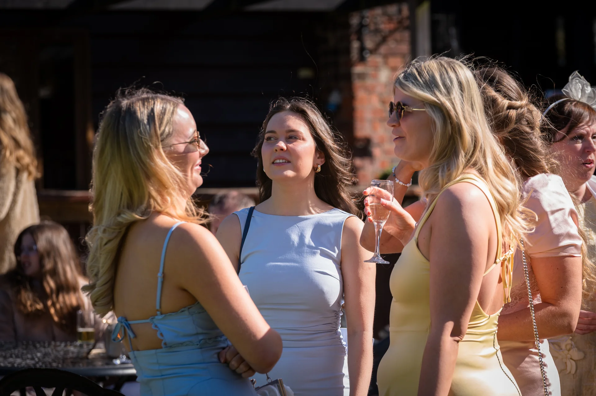 A group of female wedding guests smiling and chatting together outdoors during a sunlit drinks reception.
