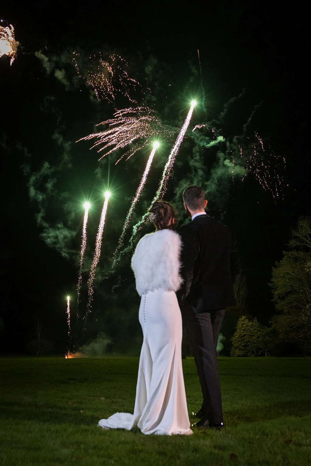 A newlywed couple stands on a lawn at night, watching a vibrant fireworks display illuminate the dark sky during their evening reception.