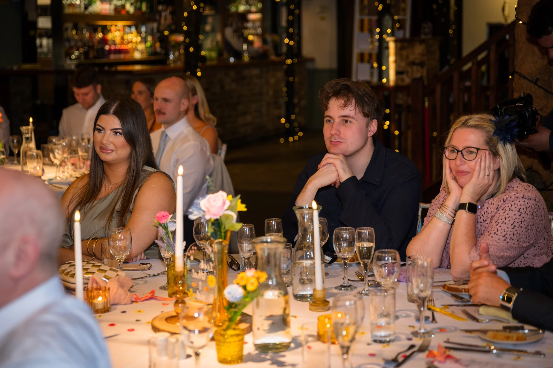 Wedding guests seated at long banquet tables during the wedding breakfast, listening intently to speeches under warm festoon lighting.