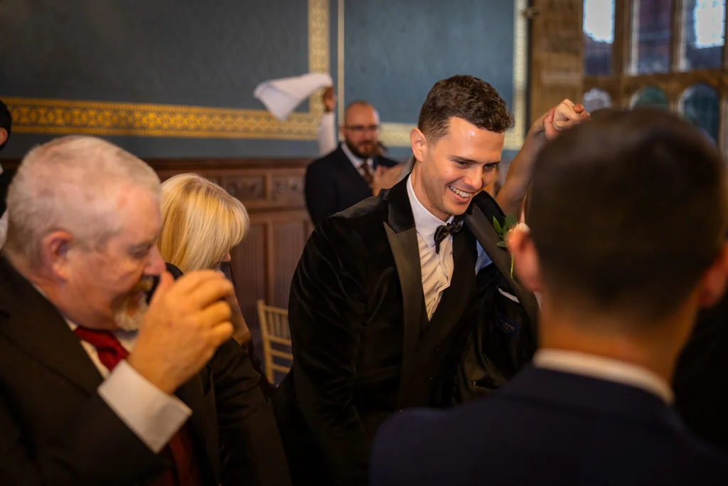 A smiling groom in a velvet tuxedo shares a joyful moment during wedding speeches while guests laugh around the table.
