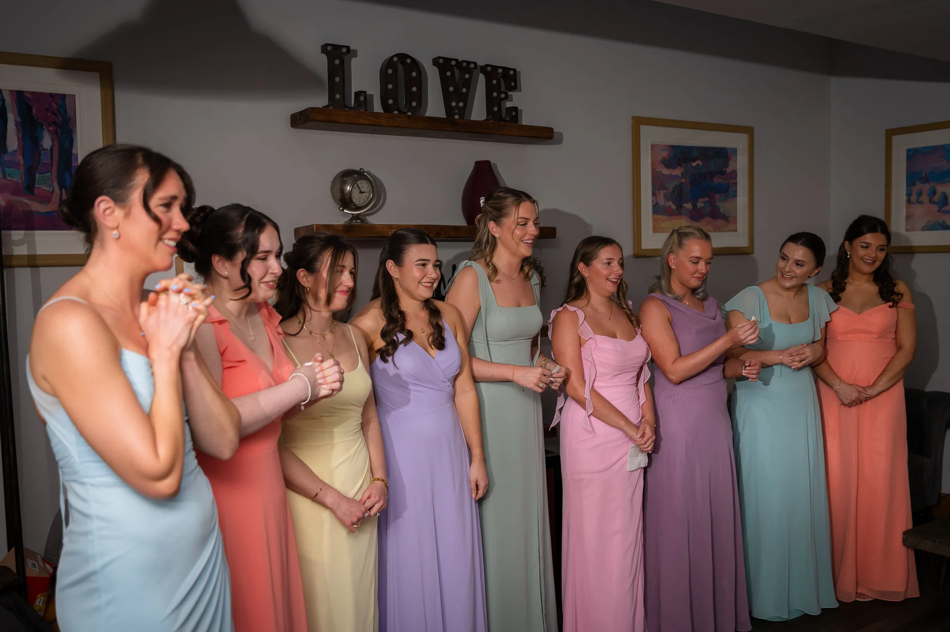 A line of smiling bridesmaids in mismatched pastel dresses stand together indoors, holding small items and looking towards the right of the frame.