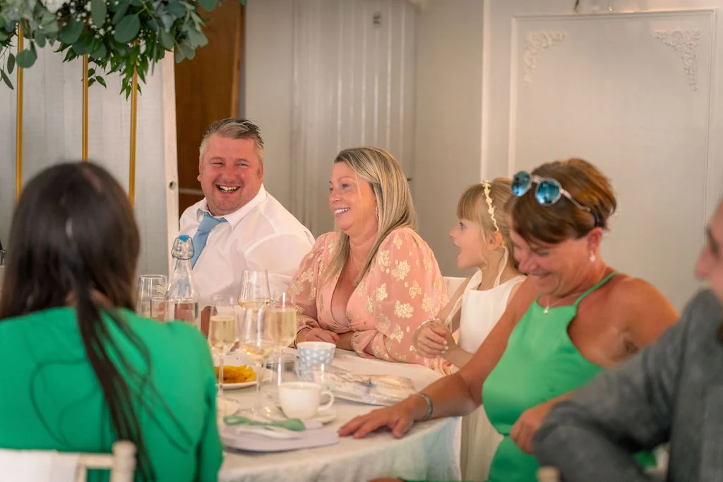 Wedding guests laugh joyfully during speeches at a brightly lit reception table adorned with wine glasses and hanging greenery.