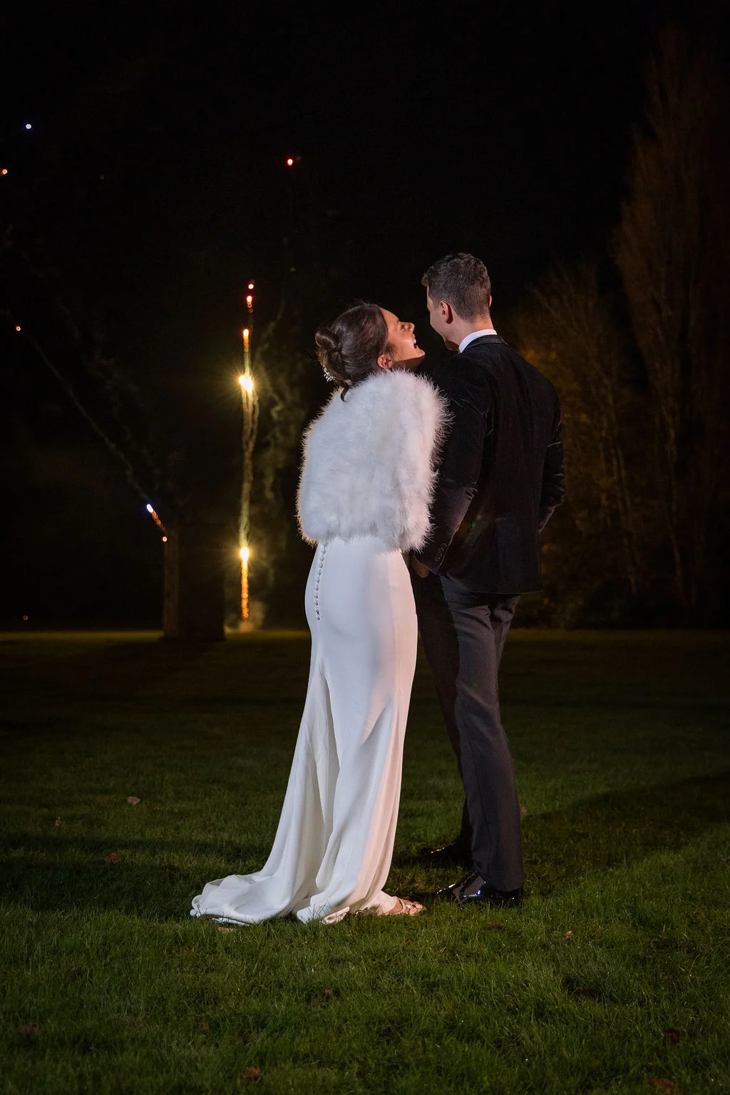 A newlywed couple stands outdoors at night on a grassy lawn, laughing together under a dark sky while watching distant fireworks.
