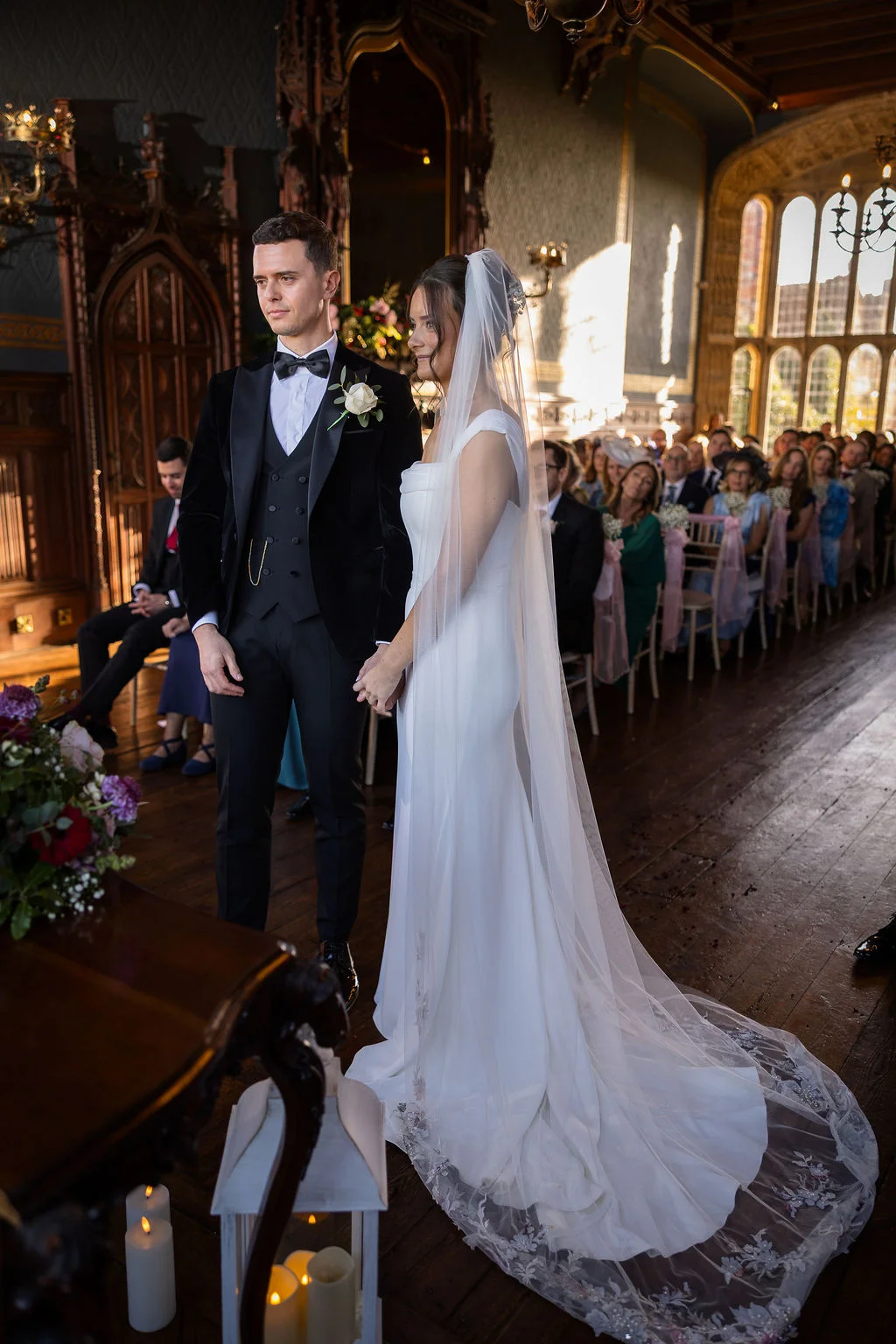 A newlywed couple stands together during their civil ceremony inside a grand, panelled historic hall illuminated by warm natural side light.