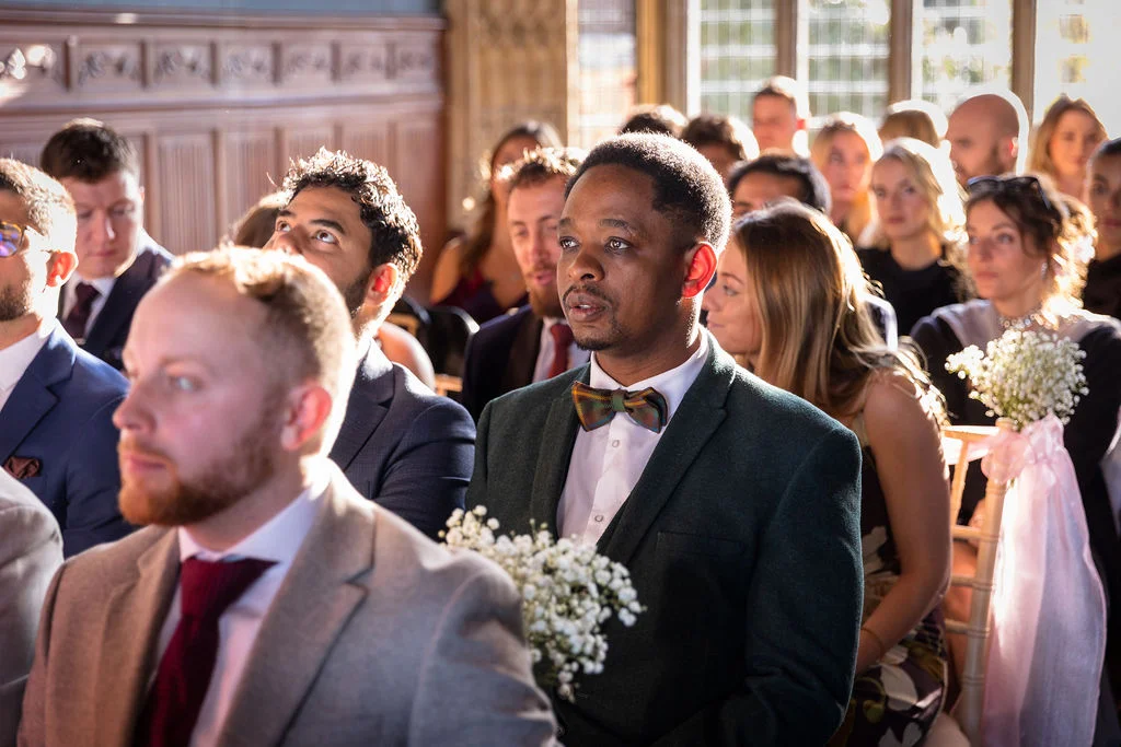 Wedding guests attentively watch the ceremony inside a historic hall with wood-panelled walls and large windows.