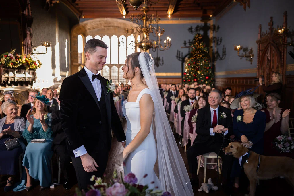 A newlywed couple smiling lovingly at each other during their winter castle ceremony while guests applaud and a dog wearing a bow tie looks on from the aisle.
