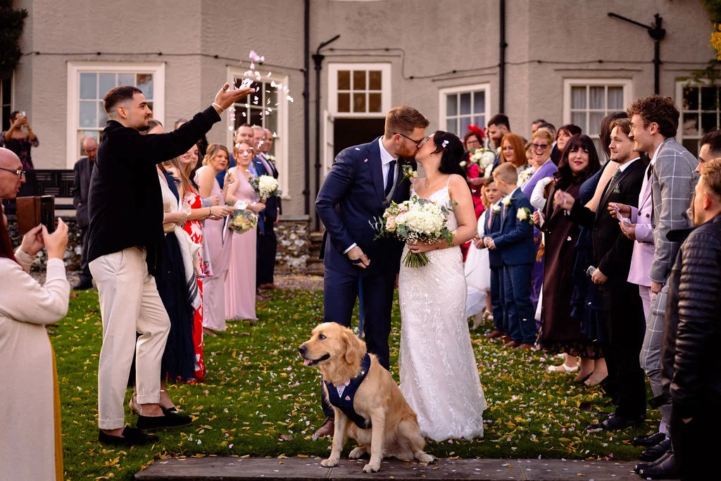 A newlywed couple share their first kiss as husband and wife amidst a shower of confetti, accompanied by their golden retriever sitting happily in front of them on the lawn outside a country house.