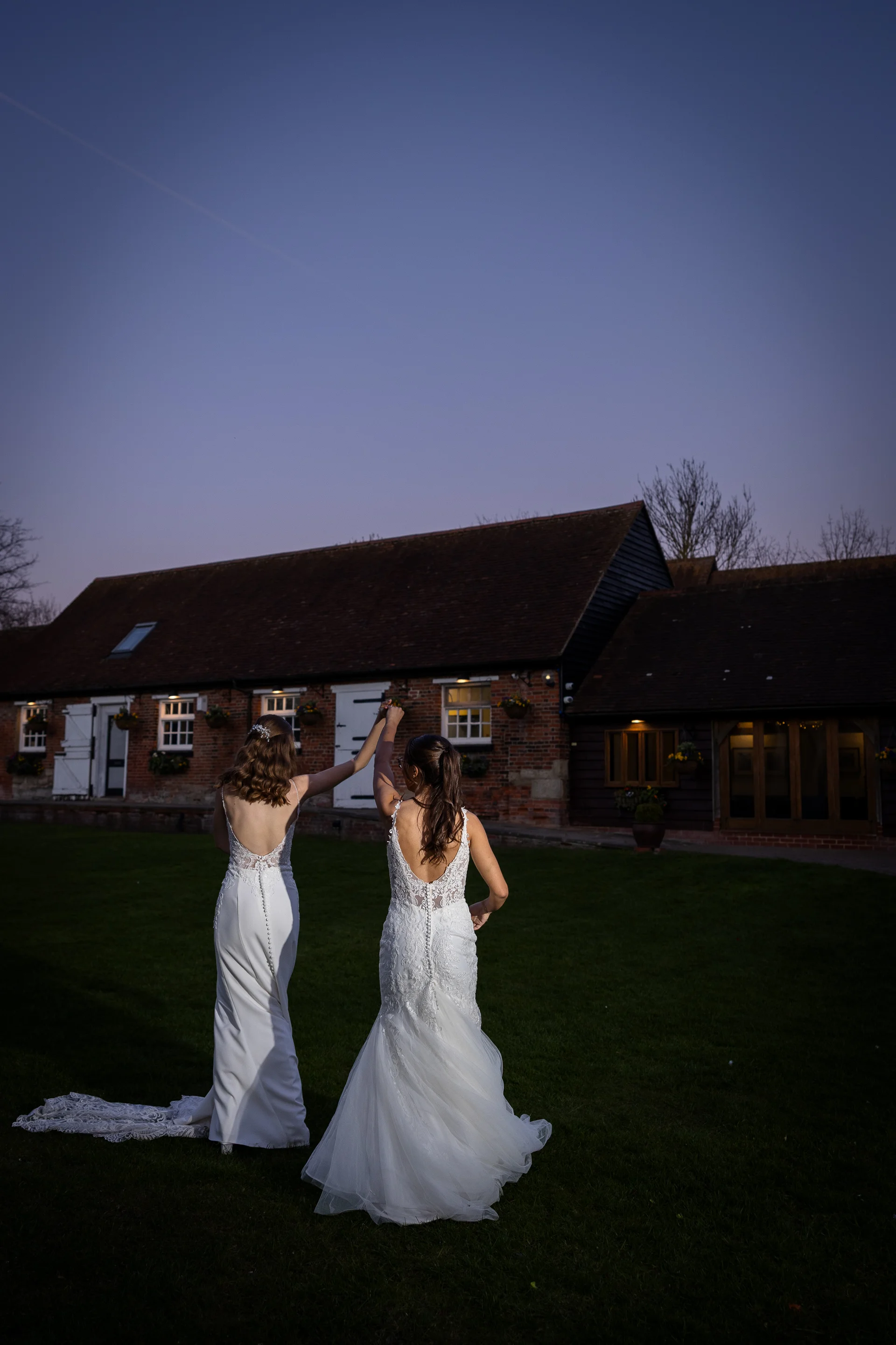 Two brides in white lace wedding dresses holding hands and walking together on a lawn outside a rustic brick barn during the blue hour.