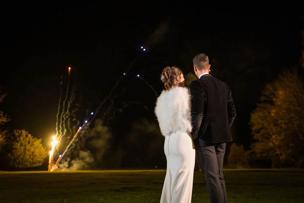 Newlyweds stand outside on the grass at night, watching a vibrant fireworks display light up the sky during their evening celebration.