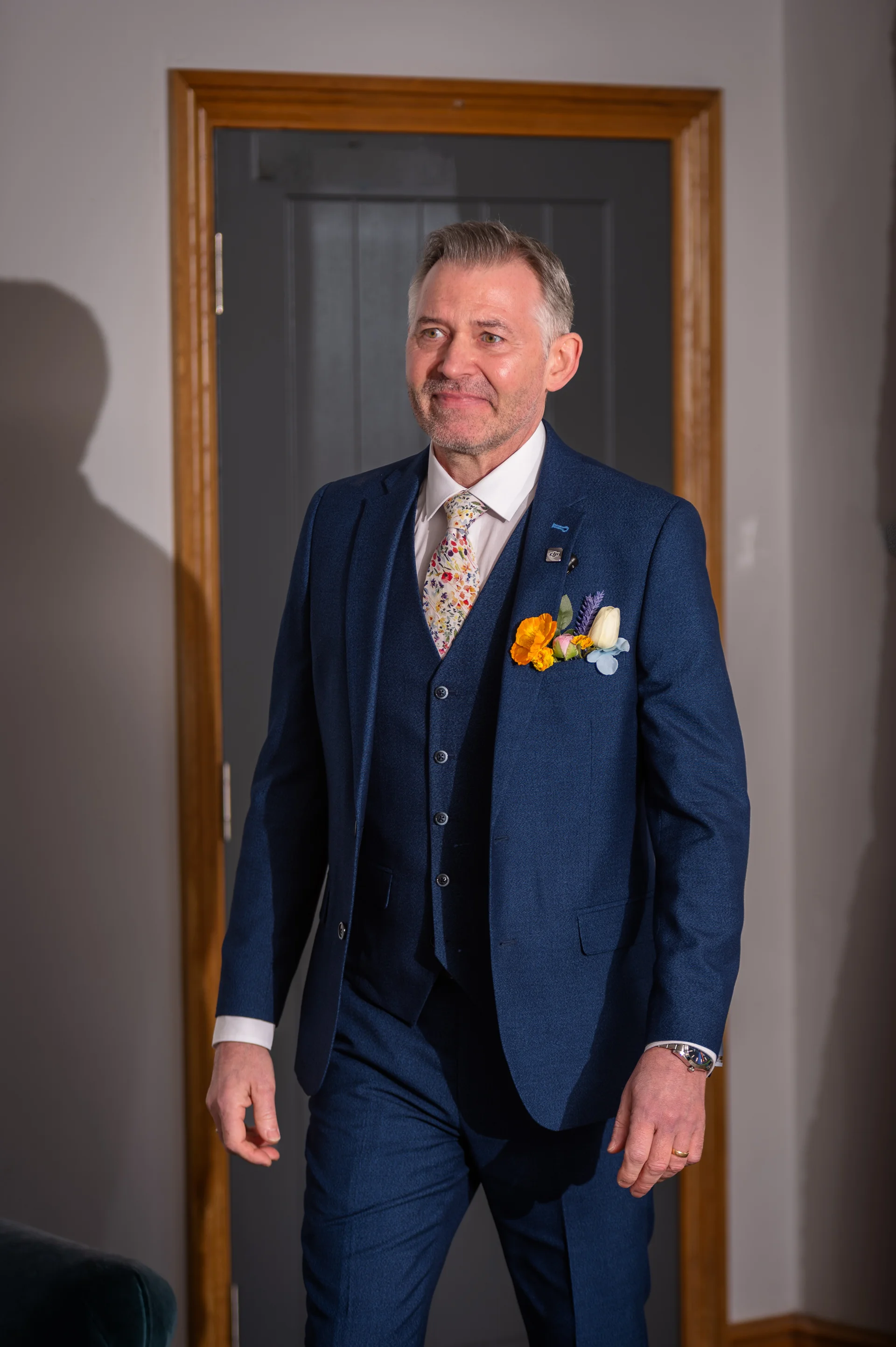 A smiling groom wearing a smart blue three piece suit stands framed in a doorway with a floral buttonhole pinned to his lapel.