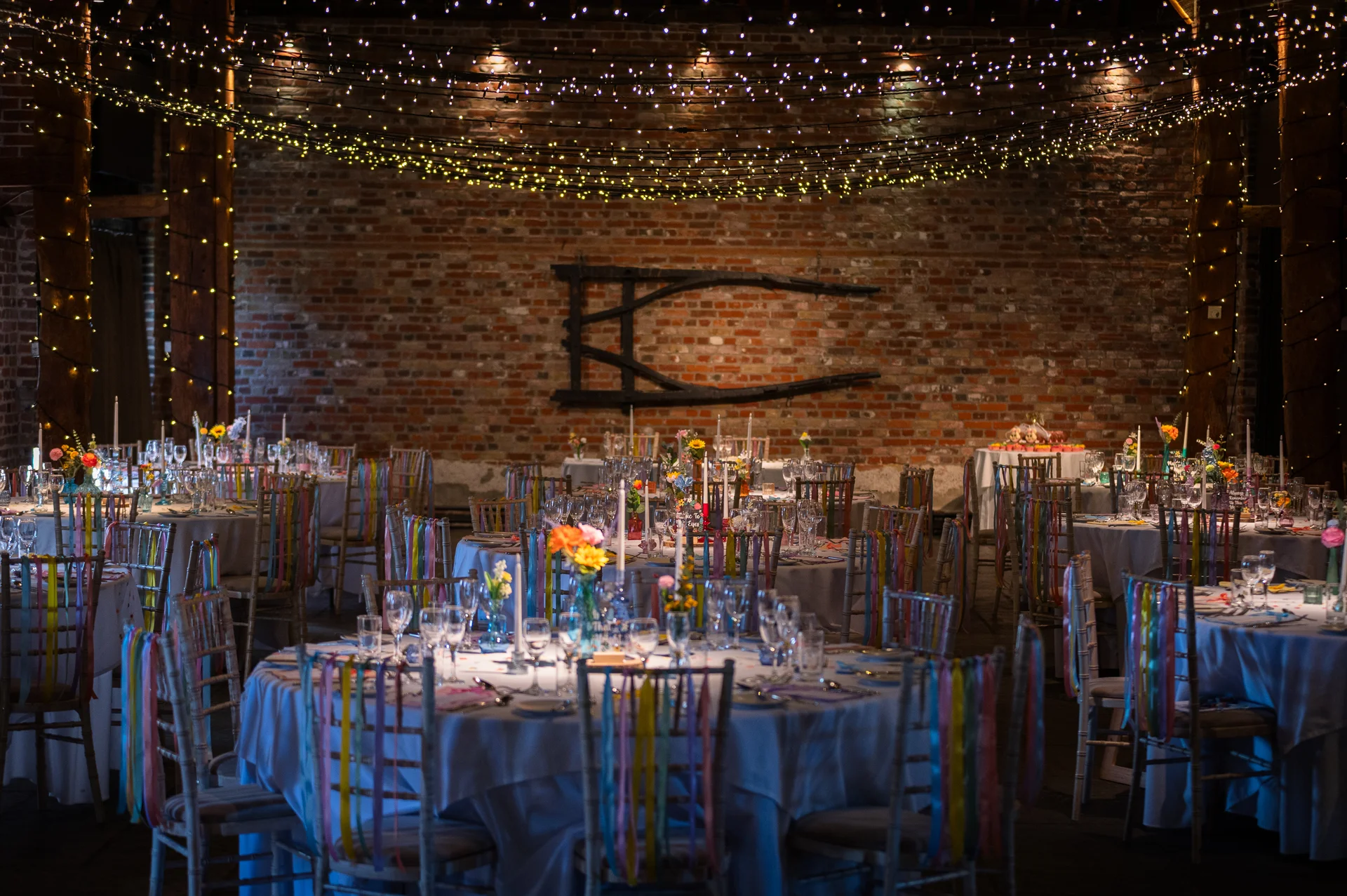 A rustic brick barn wedding reception venue features round tables beautifully set with white linens, colourful hanging ribbons on chairs, and a sparkling fairy light canopy overhead.