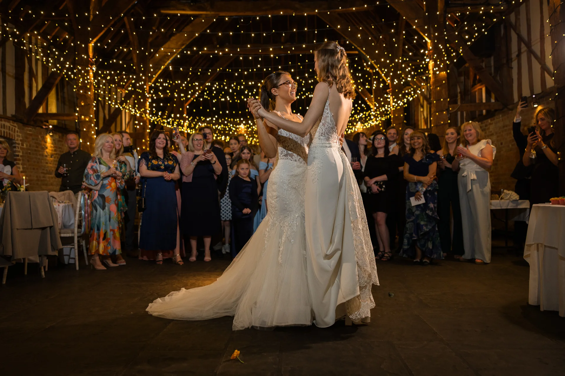 Two brides share their first dance surrounded by family and friends beneath a stunning canopy of fairy lights inside a rustic barn venue.