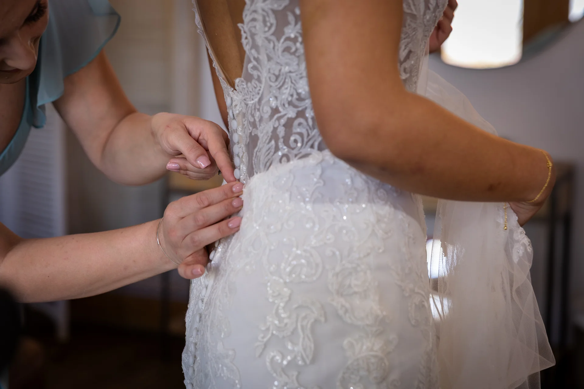 A bridesmaid carefully fastens the delicate fabric buttons down the back of a lace wedding gown during morning bridal preparations.
