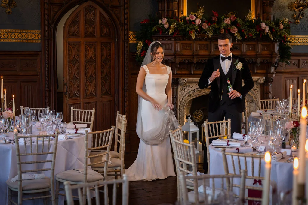 A newlywed couple enter a grand, wood-panelled dining room decorated for a winter wedding reception with candlelit tables and a festive mantelpiece.