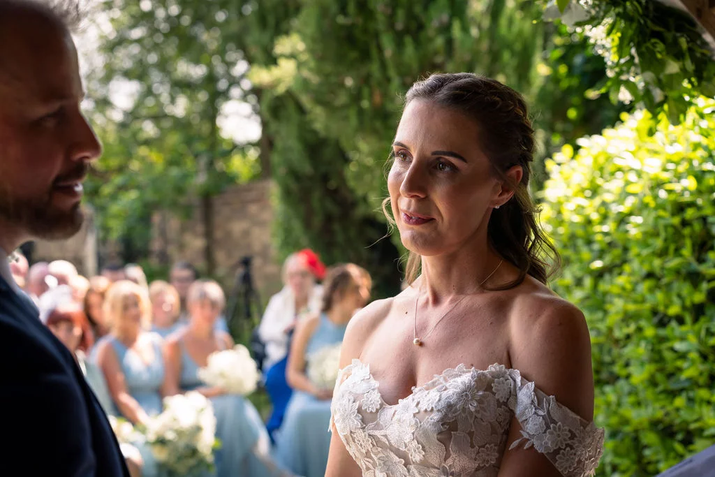 An emotional bride looks lovingly towards the groom during an outdoor ceremony, framed by lush greenery and watched by seated guests.