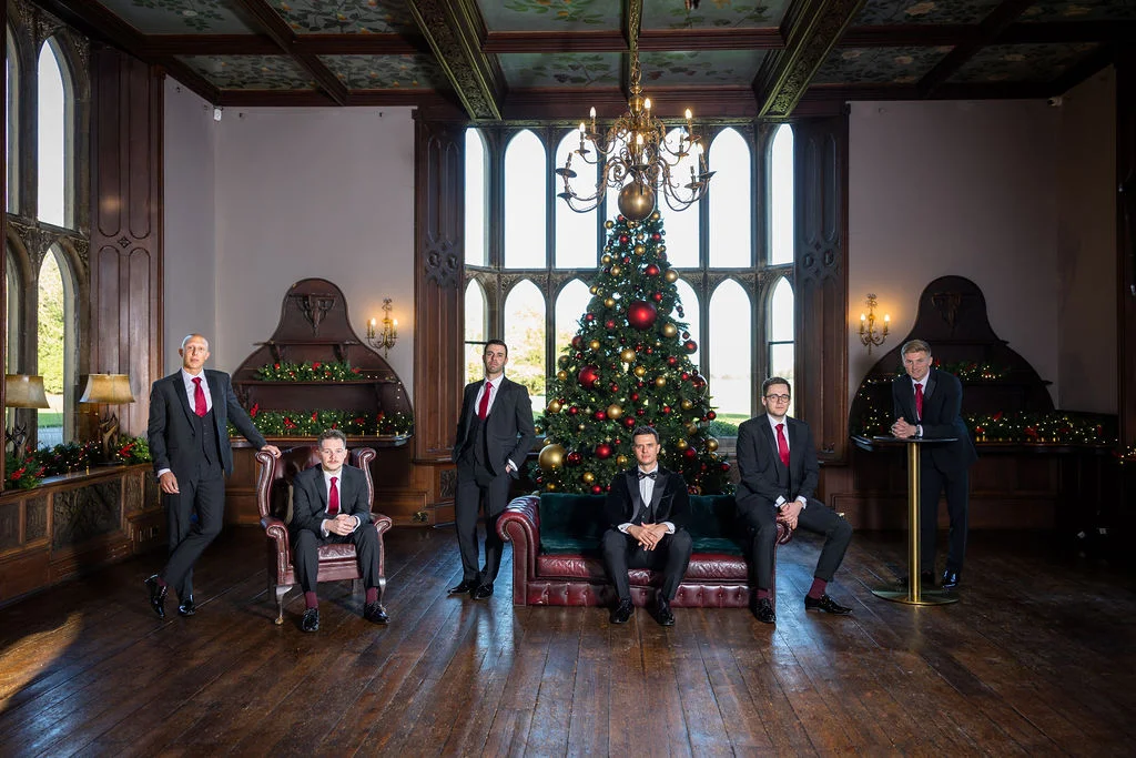 A stylish group of groomsmen pose for a portrait inside a grand wood-panelled stately home decorated for a Christmas wedding, complete with a decorated tree.