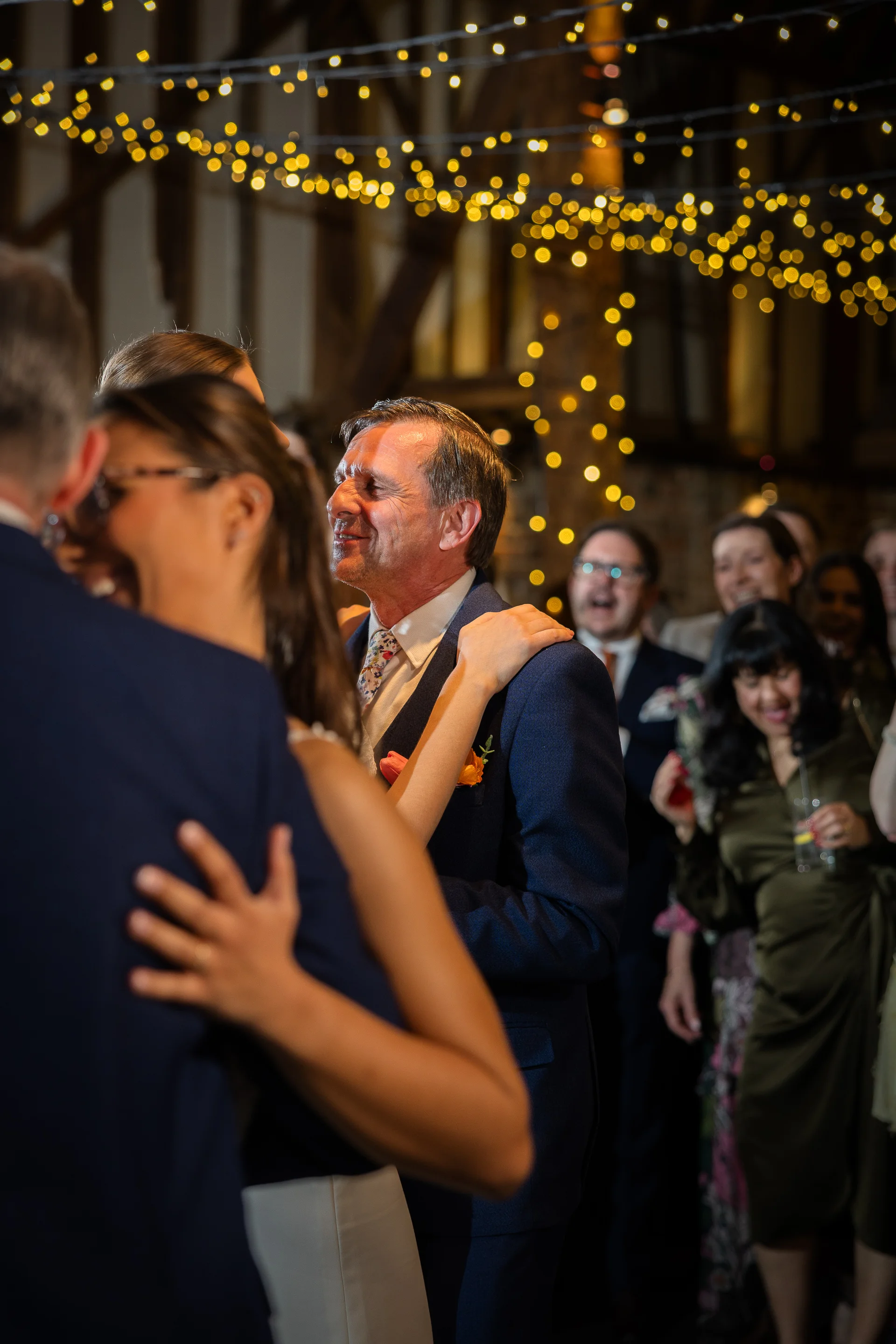 A smiling couple dances closely together on a packed dance floor under a warm canopy of twinkling fairy lights in a rustic barn.