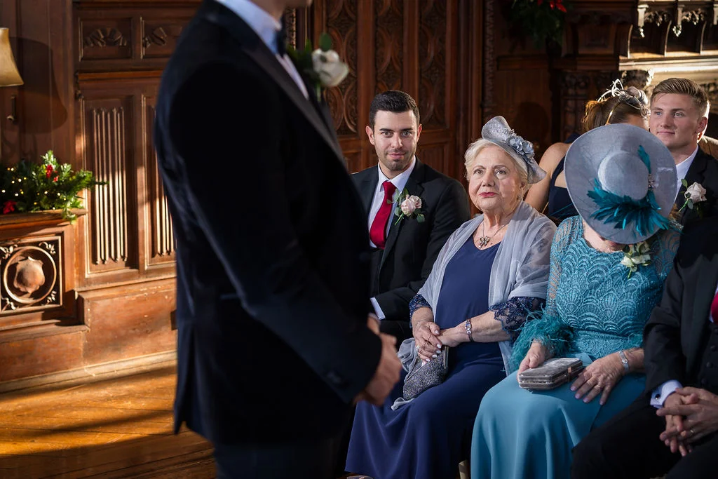 An over-the-shoulder shot captures guests watching a ceremony inside a wood-panelled room, featuring an older woman in a navy dress and grey fascinator.