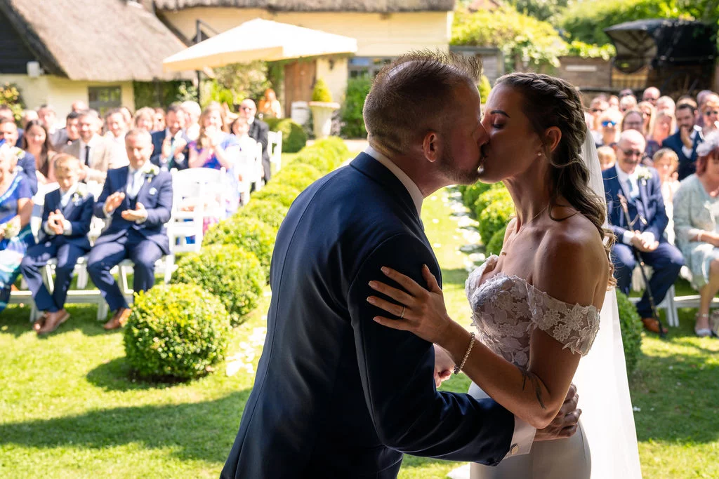 A newlywed bride and groom share their first kiss during an outdoor garden ceremony while seated guests applaud in the background.