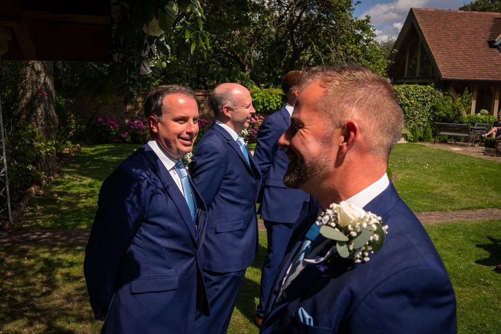 A groom and his groomsmen dressed in navy suits stand in a sunny garden awaiting the wedding ceremony.
