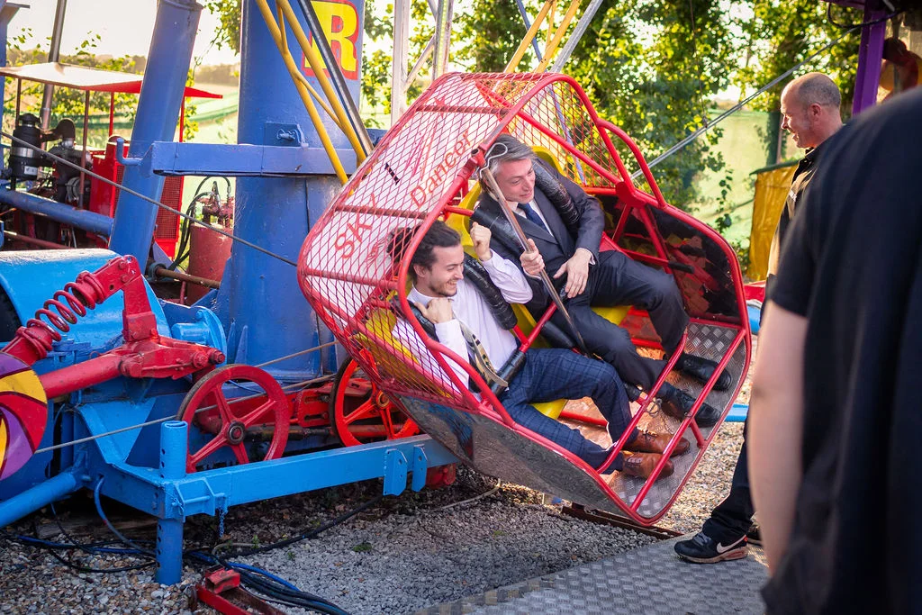 Two grooms laughing joyfully while riding a brightly coloured vintage fairground swing ride during their festival-themed wedding celebration.