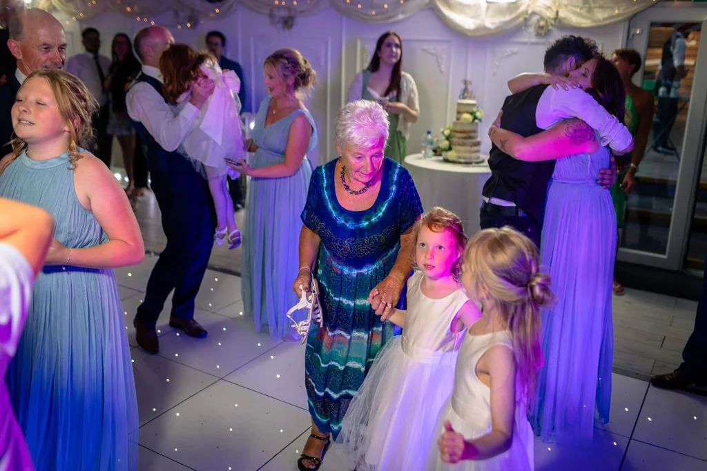 A joyous evening wedding reception scene on an illuminated dance floor, featuring family members and bridesmaids celebrating together under purple lighting.