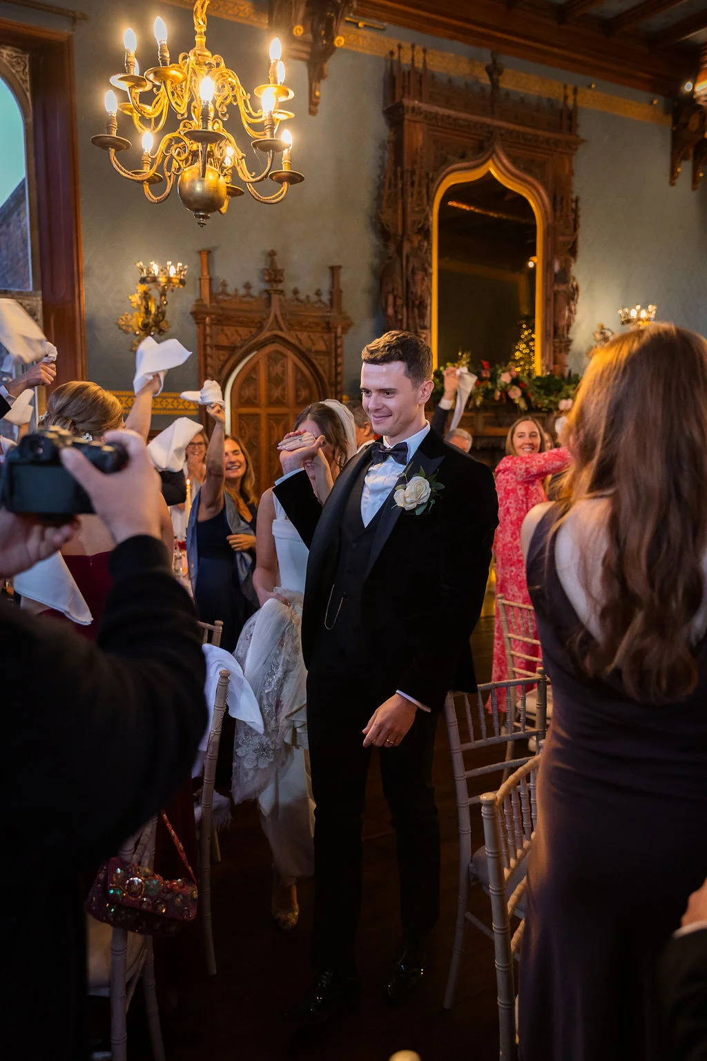 A groom in a velvet tuxedo smiles as he walks into a grand reception room while guests wave napkins and cheer.