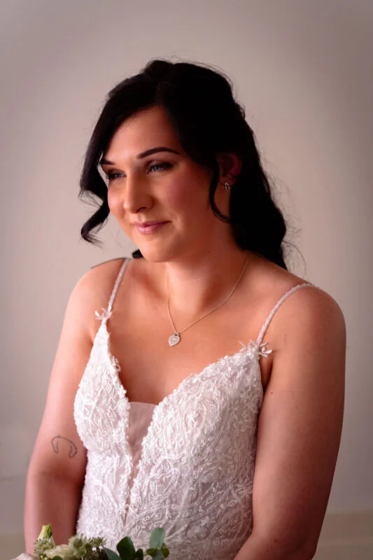 A stunning portrait of a bride wearing a lace wedding dress with spaghetti straps, holding a delicate bouquet while gazing softly to the side.