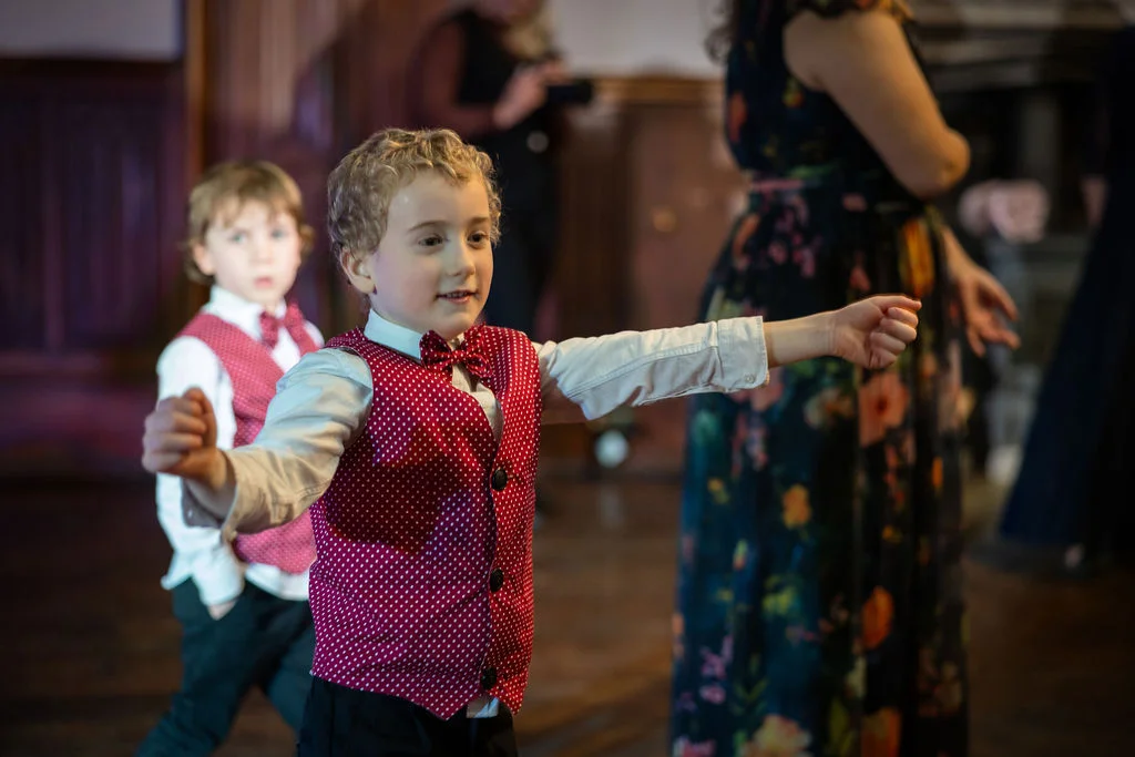 A young page boy in a red polka-dot waistcoat dances joyfully at a wedding reception while another child watches in the background.