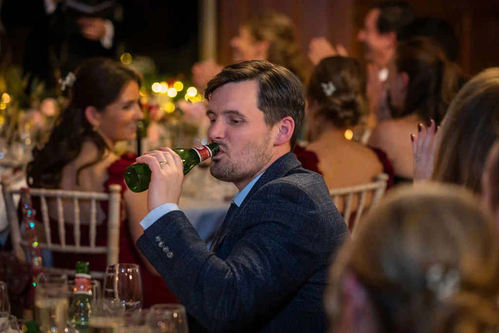 A wedding guest enjoys a drink from a glass bottle during the reception speeches while seated at a festive banquet table.