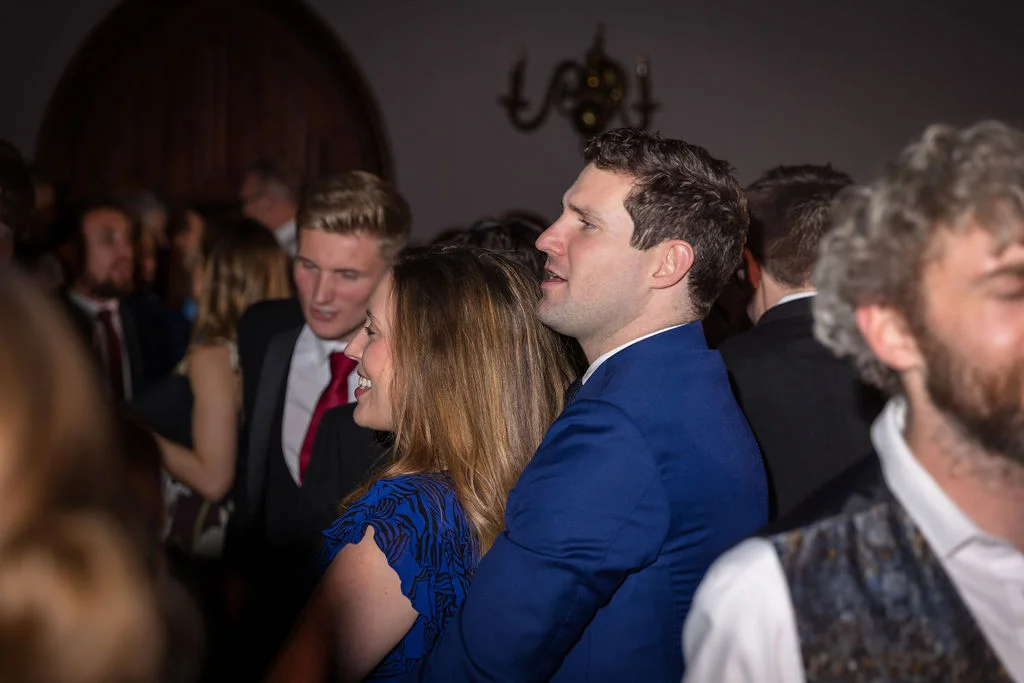 A man in a blue suit and a woman in a blue dress socialising among a crowd of guests during an evening wedding reception.