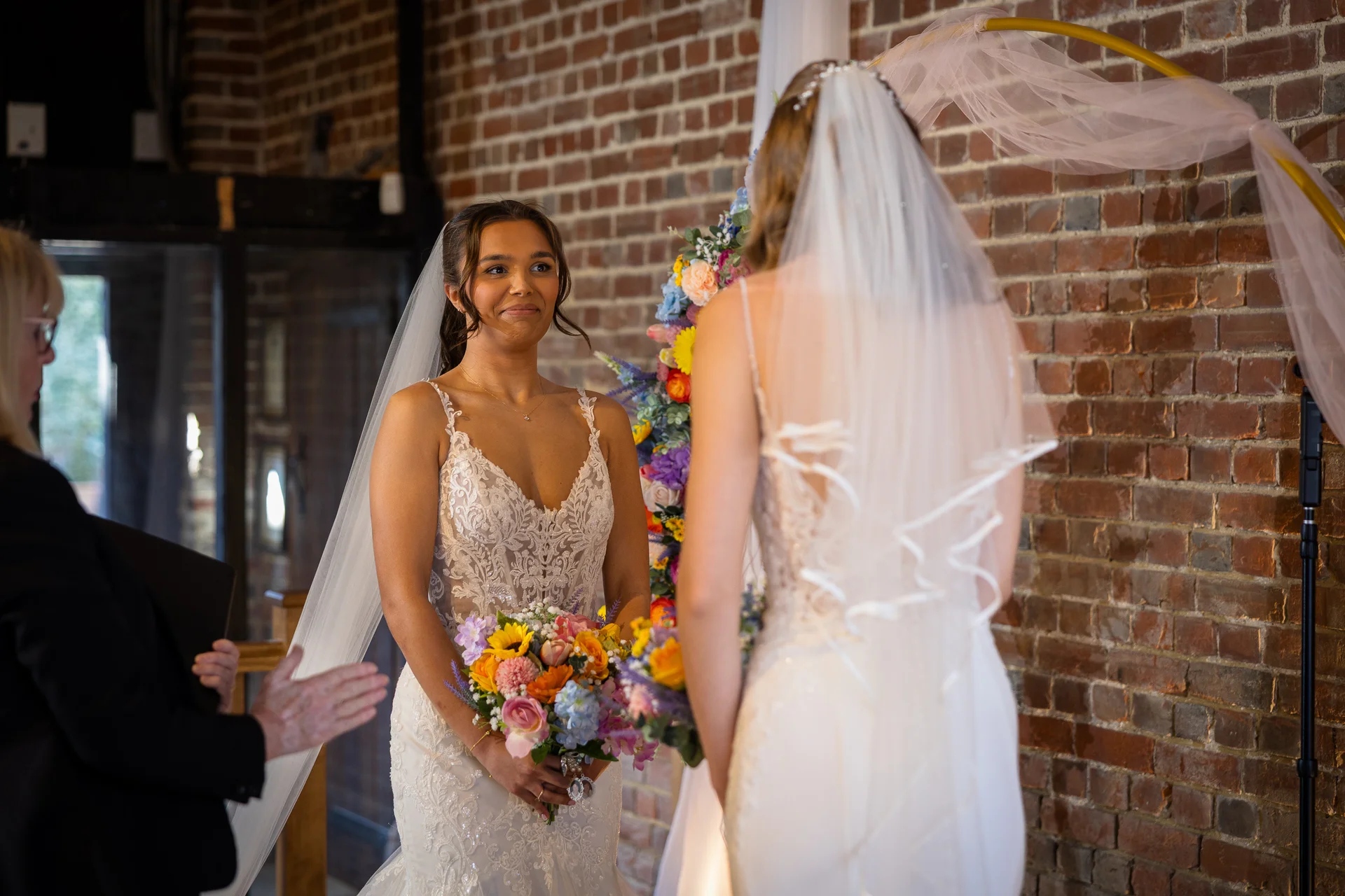 Two brides exchange emotional glances during their wedding ceremony in front of a rustic exposed brick wall and a colourful floral arch.
