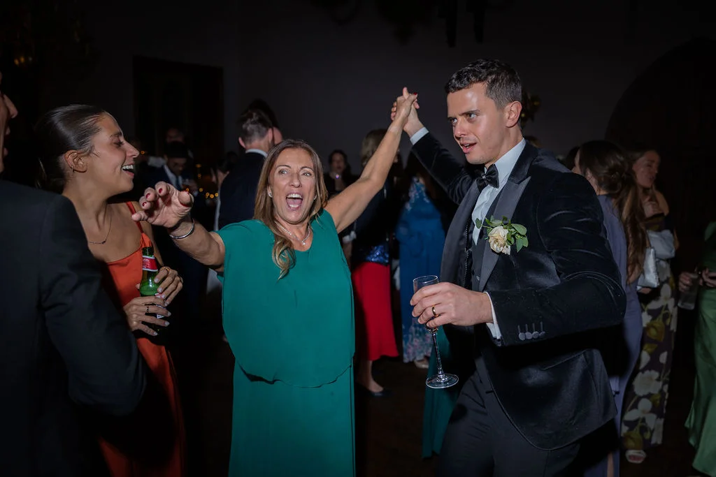 A groom in a black tuxedo and a joyful guest in a green dress dance enthusiastically together holding hands during the evening wedding reception.