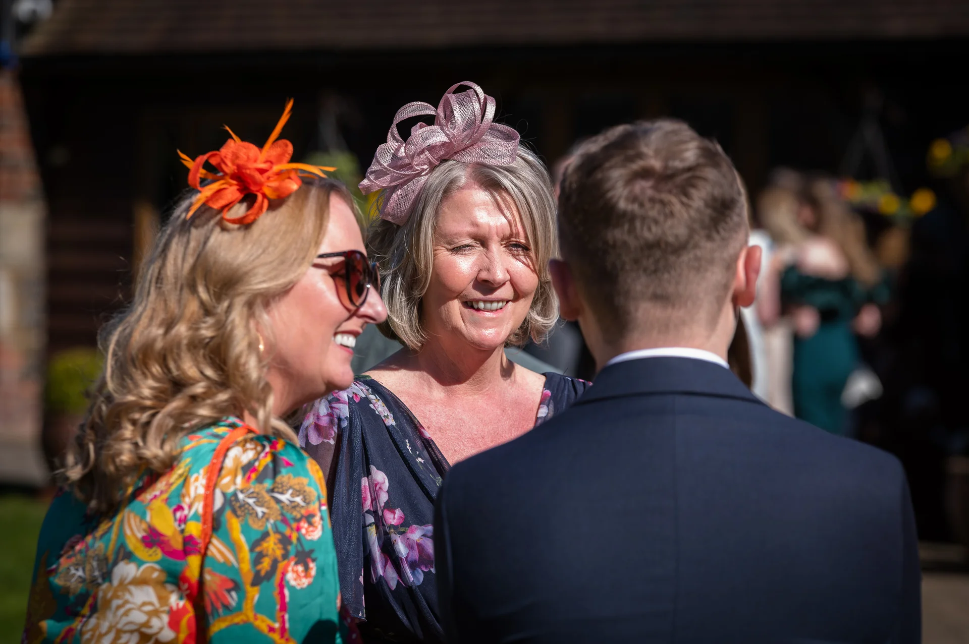 Two smiling female wedding guests wearing colourful floral outfits and fascinators chat with a guest in a dark suit outdoors in the sunshine.