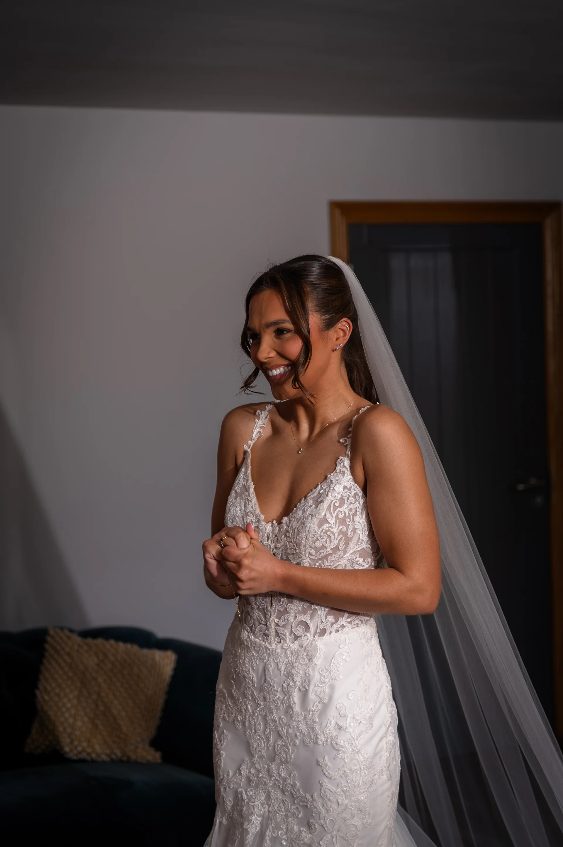 A smiling bride wearing a lace v-neck wedding dress and a veil stands indoors during morning preparations, laughing joyfully with her hands clasped.