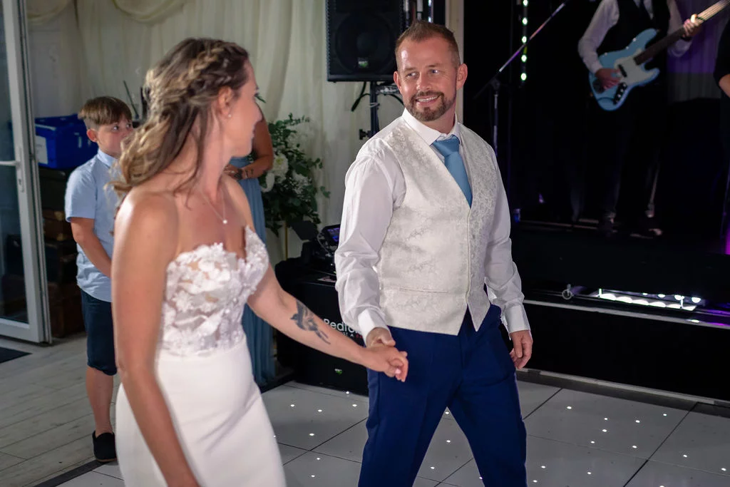 The bride and groom share a joyful moment dancing on an illuminated dance floor during their evening reception, with a live band performing in the background.