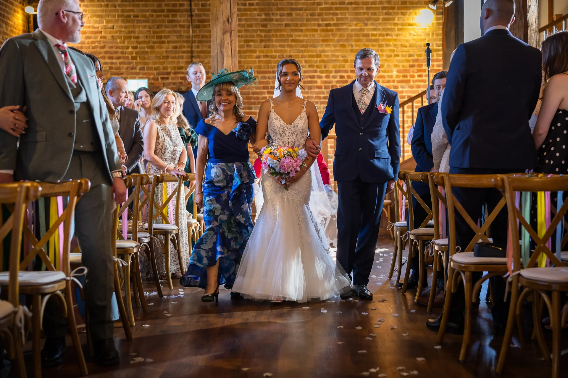 The bride walks down the aisle of a rustic brick barn accompanied by her mother and father, holding a colourful bouquet.