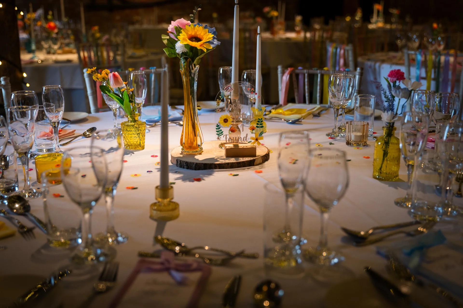 An elegantly set round wedding table features taper candles, bright sunflowers in amber bud vases, glassware, and whimsical floral table numbers.