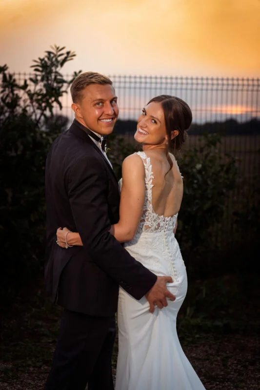 A bride and groom embracing in front of a fence at sunset.