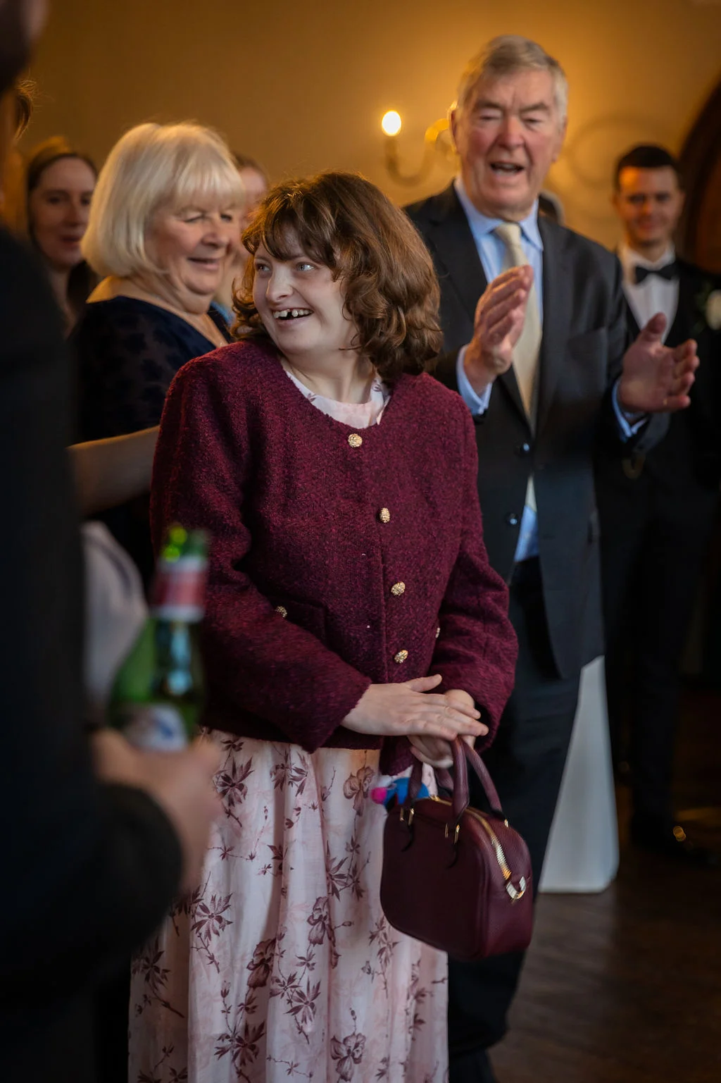 A joyful wedding guest in a burgundy jacket and floral dress smiles warmly while holding a matching handbag during the reception speeches.