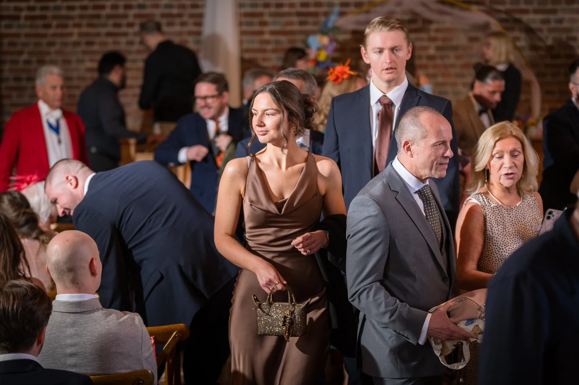 A candid group of well-dressed wedding guests mingling in a rustic barn venue with exposed brick walls.