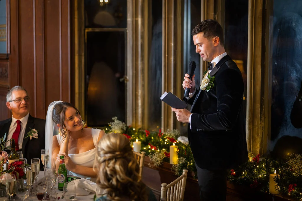 A groom in a black velvet tuxedo delivers his speech while holding a microphone and notes, looking down lovingly at his bride who smiles back from the candlelit head table.