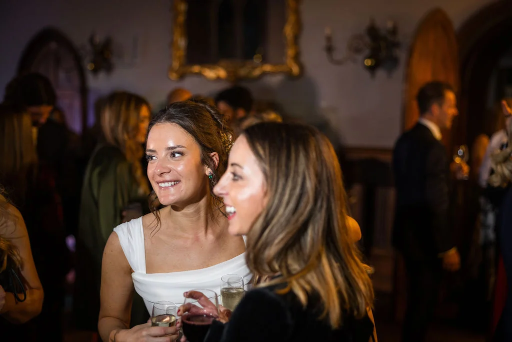 A joyful group of wedding guests mingle and laugh together during a lively evening drinks reception inside a historic venue.