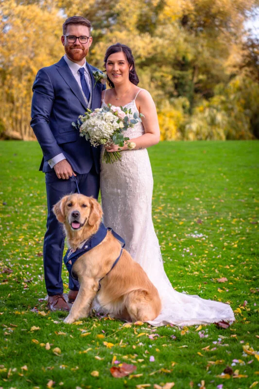 A newlywed couple poses outdoors on a grassy lawn during their autumn wedding, holding their golden retriever who wears a blue collar.