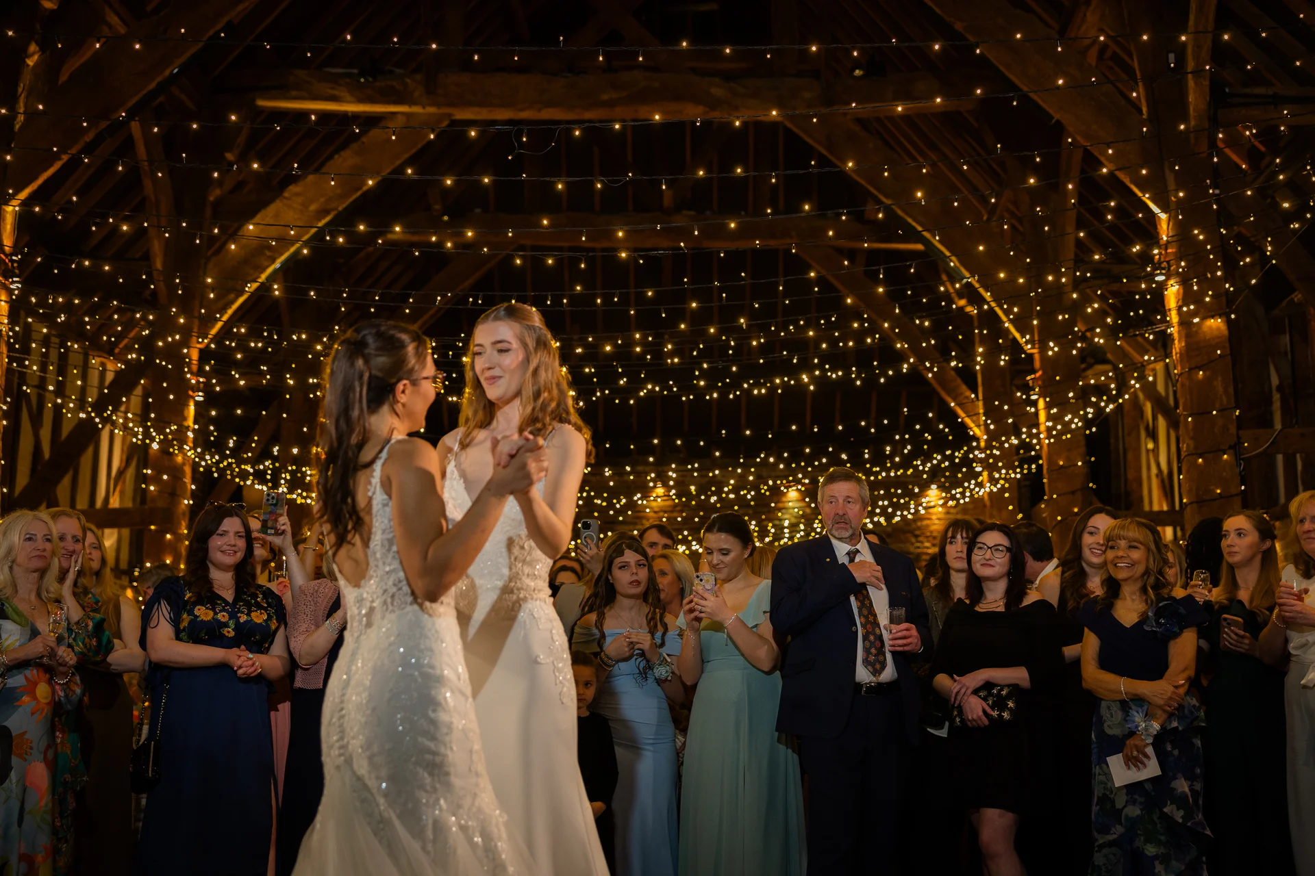 Two brides share their first dance under a magnificent canopy of fairy lights inside a rustic wooden barn surrounded by watching guests.