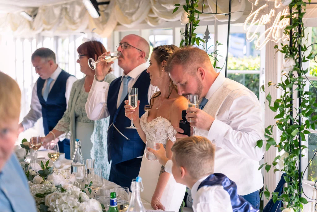 The bride and groom stand at the top table during the wedding breakfast, sharing a celebratory drink with guests in a light-filled marquee adorned with floral arrangements.