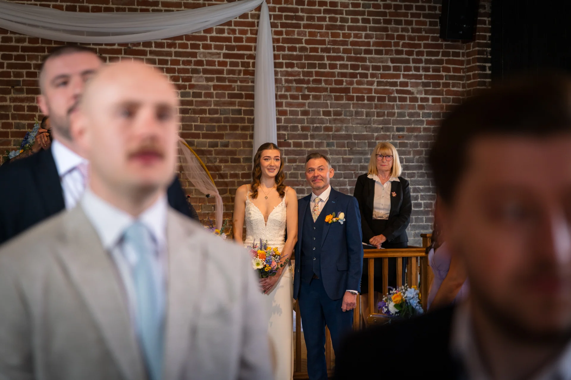 A smiling bride holding a colourful bouquet and a groom in a blue suit stand together during an indoor ceremony, framed by wedding guests in the foreground.