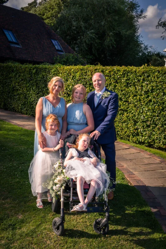 A family of five posing for a portrait outdoors in a manicured garden during a sunny summer wedding.