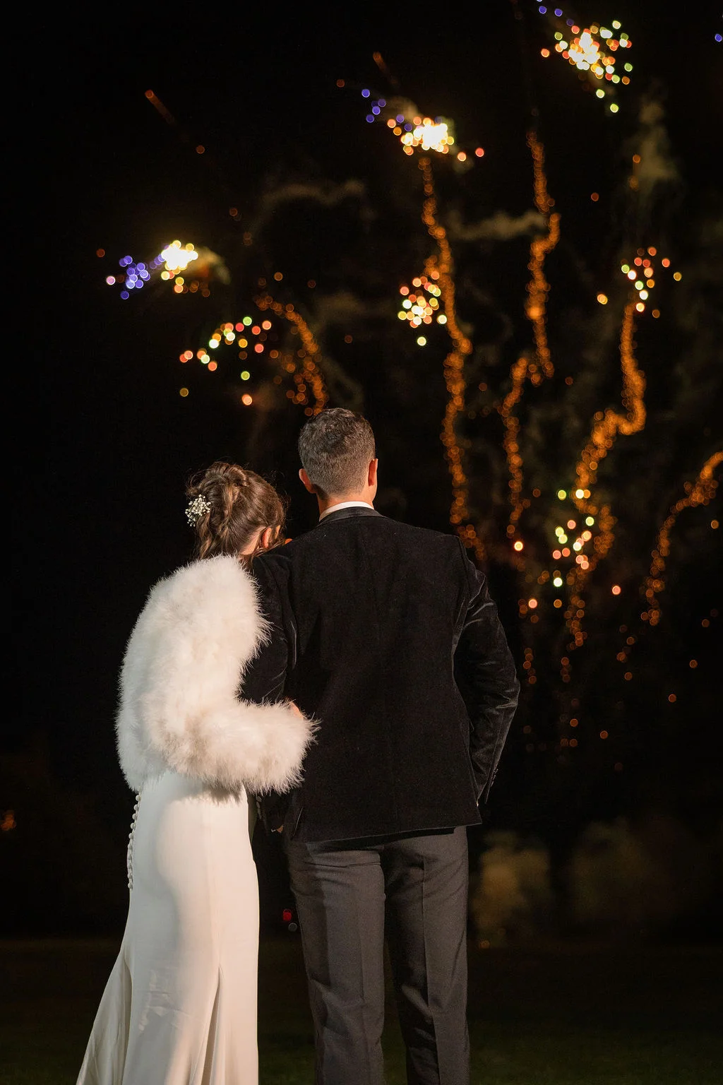A newlywed couple viewed from behind, watching a spectacular fireworks display light up the night sky during their winter wedding reception.